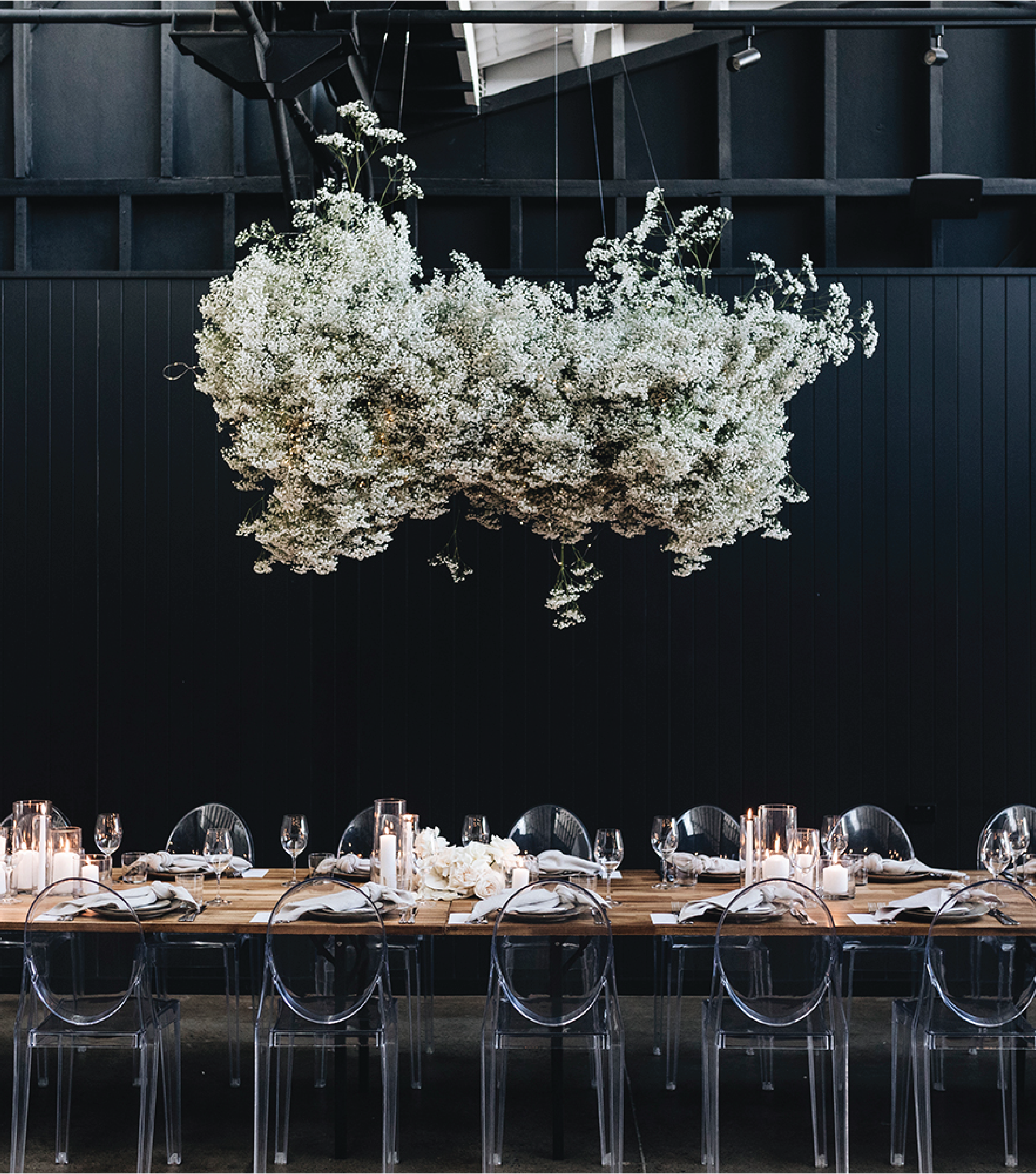 Black room with a cloud of flowers hanging from the ceiling. Clear chairs around a table.
