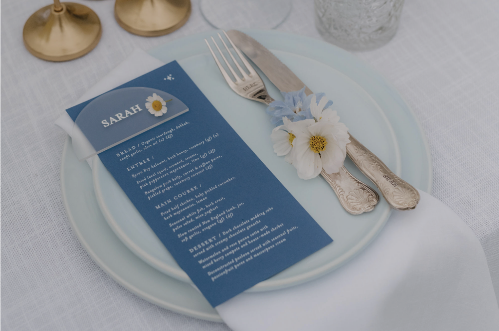 Blue menu and acrylic place card placed on top of a blue plate next to cutlery.