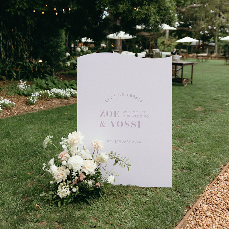 Lilac freestanding wedding welcome sign paired with an arrangement of white and purple florals.