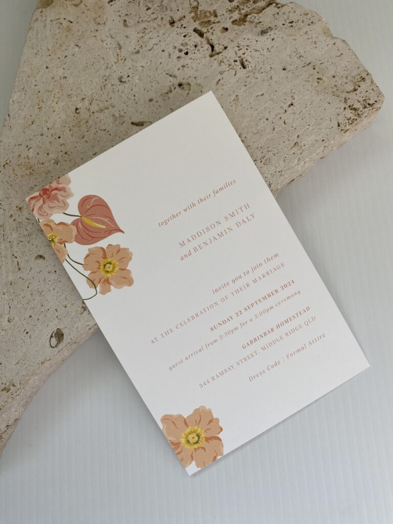 White wedding invitation with pink text and floral illustration placed on top of a decorative stone.